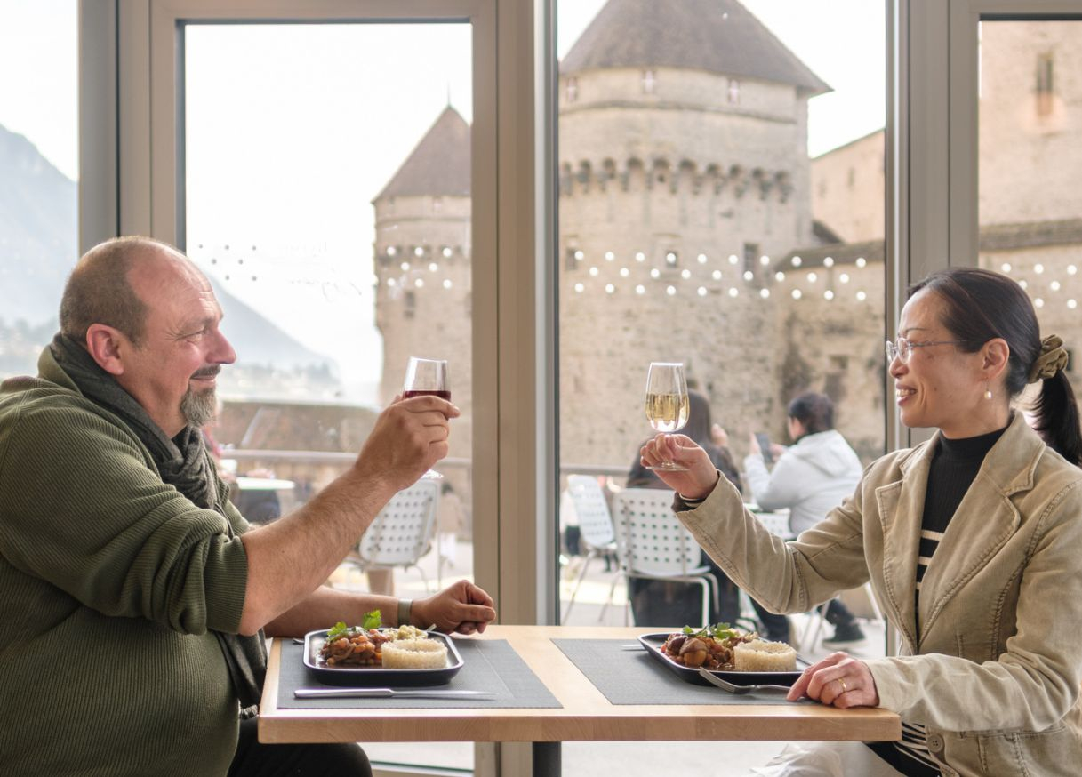 Un couple trinque et déguste le menu spécial du Café Byron, avec la silhouette du château en arrière-plan © FCC Jérôme Renevey 