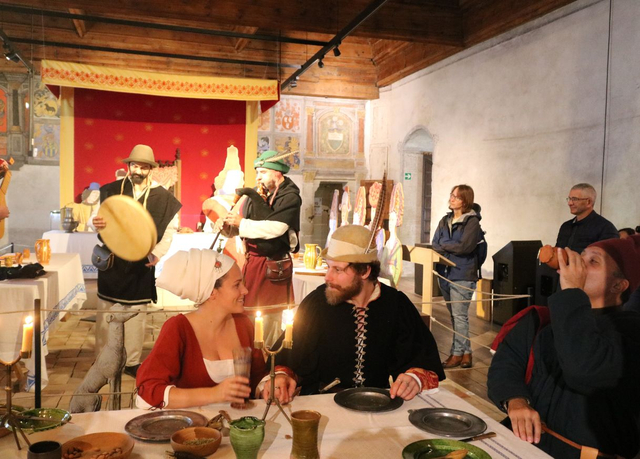 Des personnages costumés se régalent autour d'une table, avec plusieurs musiciens en arrière-plan 